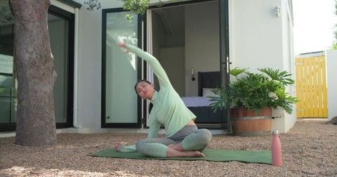 Asian woman practicing seated side stretch on backyard yoga mat in green activewear with pink bottle