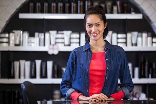 This image shows a smiling female hairdresser standing confidently at a table in a modern salon. She is wearing a denim shirt over a red shirt, with shelves of beauty products in the background. Ideal for use in articles or advertisements related to beauty services, hair care, professional profiles, or customer service in the beauty industry.