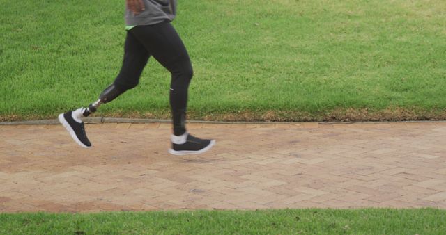 Person jogging on brick path next to green grass. Scene depicts fitness and active lifestyle of someone with a prosthetic leg. Useful for representing inclusion, adaptability, sports activities, and health.