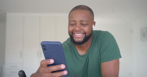 Smiling african man holding smartphone wearing earbuds and green t-shirt at home