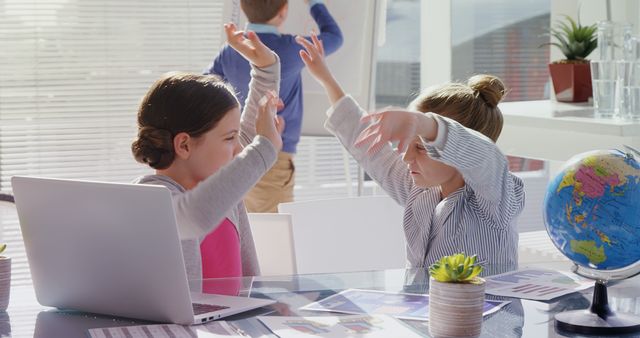 Two young schoolgirls celebrating success in a bright and airy classroom. They are smiling and putting their hands up in excitement over their achievements. A laptop, educational charts, small potted plants, and a globe are placed on the desk. A boy standing near a window is writing on the whiteboard in the background. This image can be used for educational content, cooperative learning advertisements, school promotions, or teamwork-themed materials.