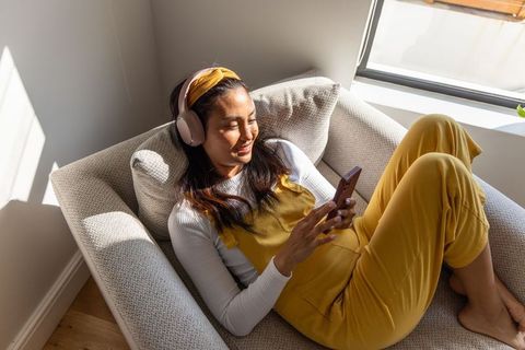 Sunlit asian woman reclining on cozy armchair wearing yellow overalls and pink headphones