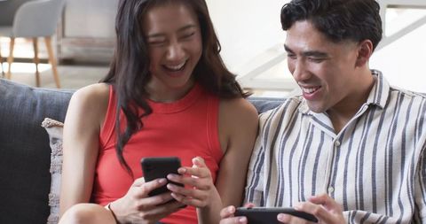 Laughing diverse couple sharing smartphones on couch in cozy modern living room