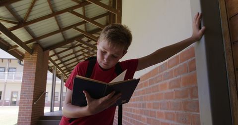 Happy schoolboy reading in school corridor leaning on wall indoors