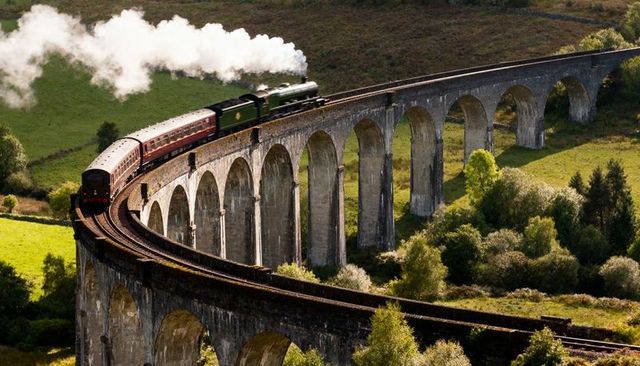 Green steam locomotive crossing curved stone viaduct with maroon coaches in scenic valley