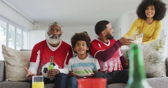 Multigenerational Black family gathered in the living room watching a football game. Grandfather holding a beer, son smiling, father reaching for snacks, and mother standing nearby with more snacks. This image captures moments of family bonding and can be used to depict sports enthusiasm, family unity, leisure activities, and togetherness. Perfect for content related to sports events, family lifestyle, and home gatherings.