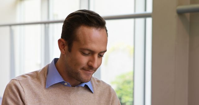 Man sitting indoors near large windows, looking thoughtful and relaxed. Perfect for conveying themes of contemplation, casual office scenarios or lifestyle moments. Useful for websites, blogs, or advertisements focused on mental well-being, casual workplace settings, or personal reflections.