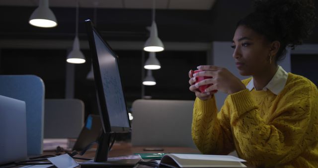 A woman wearing a yellow sweater sits in a dimly lit office, focused on a computer screen while holding a red cup of coffee. Ideal for illustrating concepts of late-night work, dedication, professional environments, and focused work.