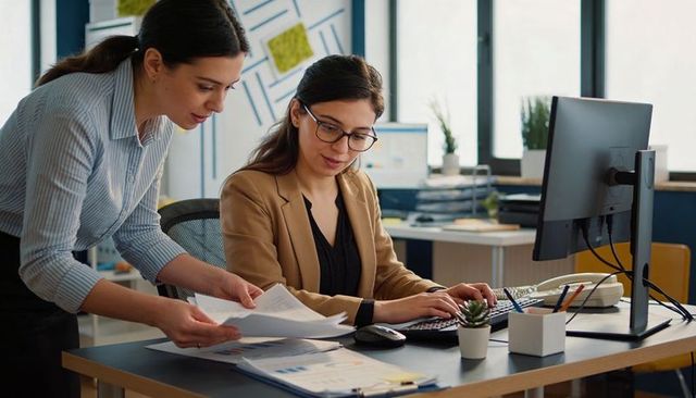 Two women collaborating at desk analyzing charts, typing on computer in bright office