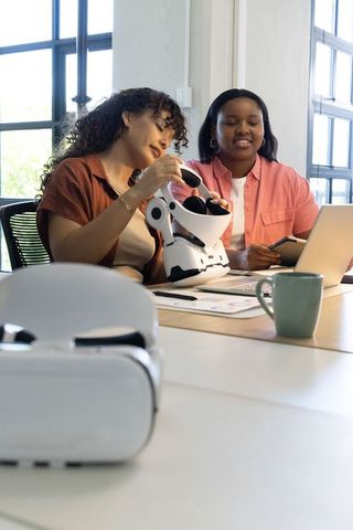 Diverse women collaborating over white vr headset in bright office using laptop and tablet