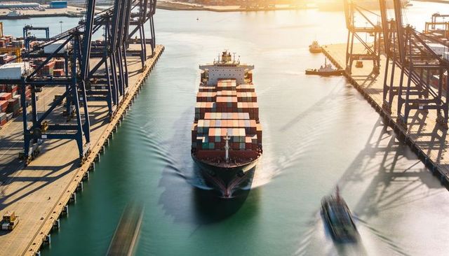 Drone aerial of container ship transiting commercial seaport with gantry cranes and tugboats