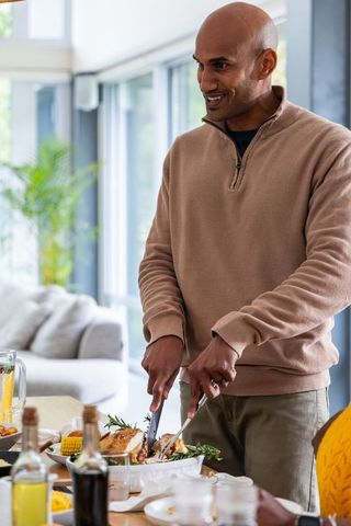 Smiling indian man carving roast poultry at bright modern open-plan dining table