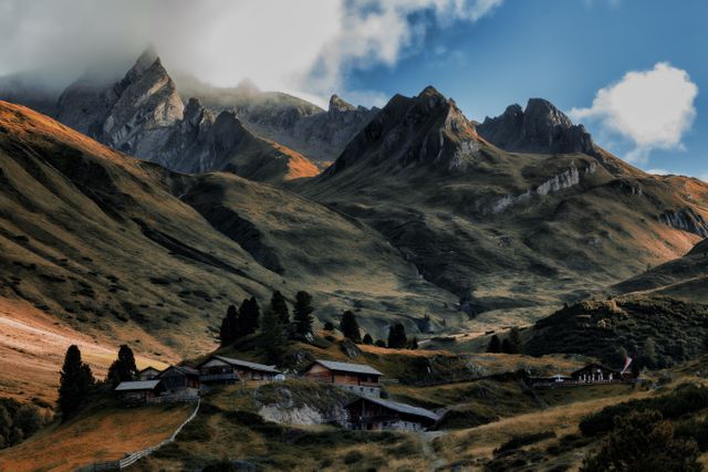 Depicting an isolated mountain village nestled in an Alpine valley, this image showcases rustic cabins set against dramatic mountain peaks under partially cloudy skies. It captures the essence of tranquility and natural beauty in an autumn setting. Ideal for use in travel magazines, tourism advertisements, and nature-themed projects to evoke a sense of peace and serene lifestyle in remote mountainous areas.