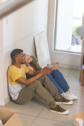 Diverse couple relaxing in new apartment holding mugs amid moving boxes and canvas