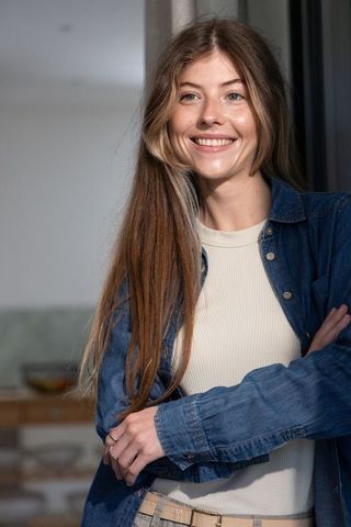 Smiling young woman leaning with crossed arms wearing denim shirt in modern home interior