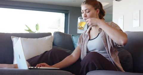 Woman sipping tea while working on laptop on modern living room sofa, remote work