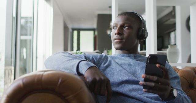 Young man sitting casually on a couch, wearing headphones and holding a smartphone. Suitable for lifestyle blogs, music app promotions, technology advertisements, and relaxat documentation videos.