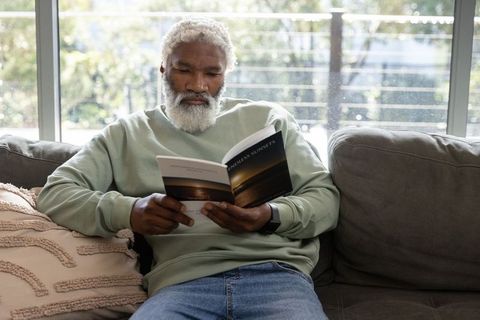 Senior african american man reading book on sofa by sunlit window, relaxing at home