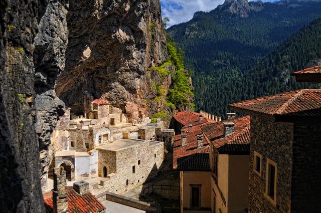 A historic stone monastery built into mountain cliffs surrounded by a lush forest. Ideal for travel brochures, destination articles, or promotion of historic and serene locations.
