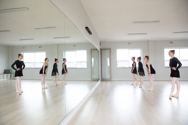 Group of Caucasian female attractive ballet dancers warming up and practicing in a bright ballet studio, stretching and looking in a mirror. Focused on their exercise, preparing for a class, talking.