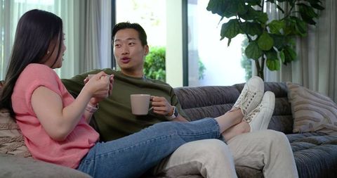 Asian couple relaxing on gray sectional sofa drinking coffee in cozy living room