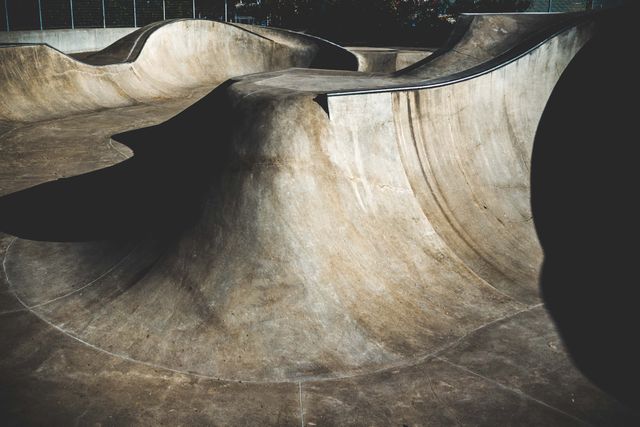 Concrete skatepark characterized by smooth curves and ramps, captured in early morning light creating dramatic shadows. Ideal for use in projects related to extreme sports, urban playgrounds, architectural design, and youth culture.