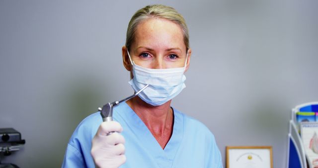 Smiling caucasian female dentist in face mask and scrubs holding suction tool at dental surgery. Dentistry, oral care, medical services, hygiene and work, unaltered.