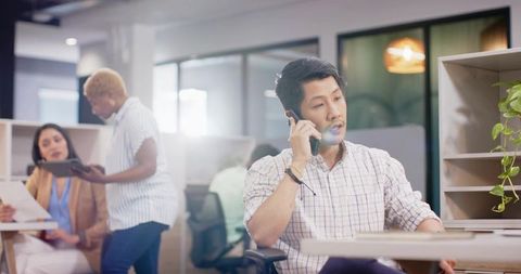 Midadult asian professional talking on smartphone in modern open-plan office teamwork scene