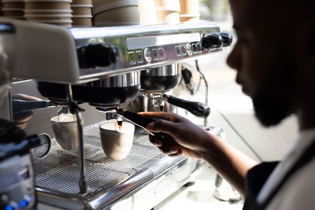 African American barista using a coffee maker to brew coffee in a cafe kitchen. Ideal for content related to cafe culture, professional baristas, coffee making techniques, and small business operations.