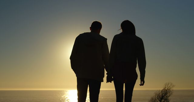 Couple standing at beach holding hands while sun sets in background. Ideal for themes of romance, relationships, and travel. Perfect for use in greeting cards, travel brochures, and love stories.