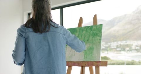 Senior woman painting green abstract canvas by window with coastal mountain view