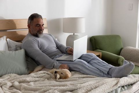 Mature man relaxing on bed with laptop while petting sleeping golden retriever puppy