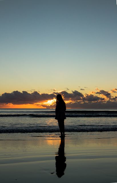 Depicts tranquil moment as silhouetted woman stands alone on beach watching sunset over ocean. Perfect for promoting travel destinations, illustrating relaxation and introspective moments, or using in wellness and mindfulness content.