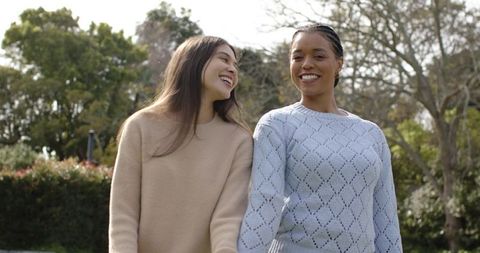 Laughing friends walking through sunlit park wearing cozy sweaters enjoying outdoor friendship