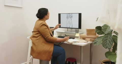 Mid-adult woman designing dress in home studio pointing at monitor with fashion sketch