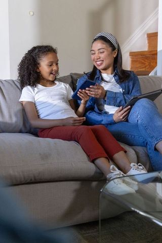 Multiracial mother and daughter sharing smartphone and tablet on cozy sofa, smiling
