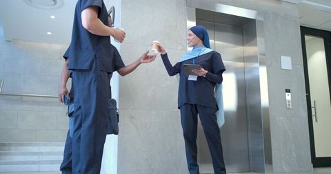 Healthcare team sharing coffee by elevator while using tablet in clinic corridor