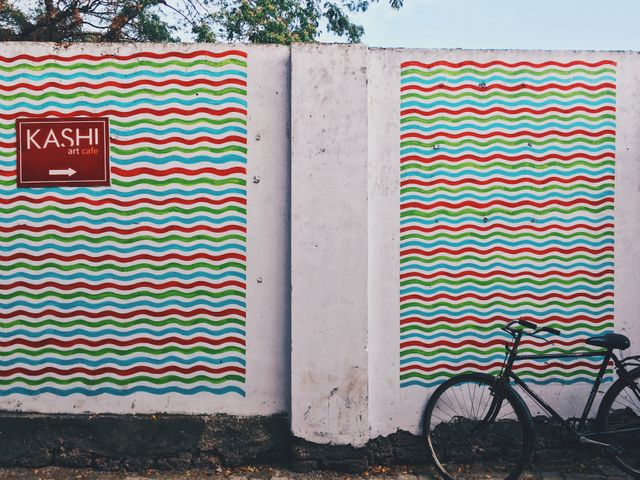 Bright and colorful patterned artwork adorns a wall, featuring a sign for Kashi Art Cafe and a parked bicycle. This vibrant and eye-catching setting is perfect for use in articles about urban art, travel, and local culture, or as inspirational decor and design concepts.