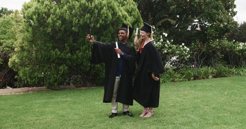 Graduates taking selfie on campus lawn wearing gowns and holding diplomas