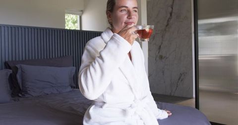 Sipping morning tea in plush white robe on bed edge in contemporary marble bedroom