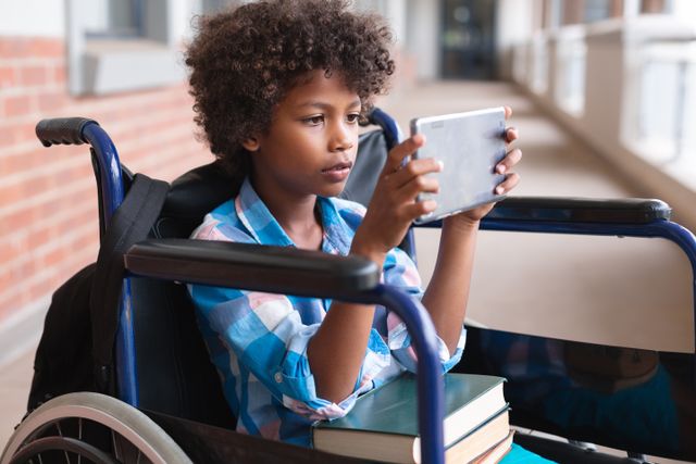 Young African American boy sitting in a wheelchair using a digital tablet in a school corridor. He has books on his lap, indicating a focus on education and learning. This image can be used for educational materials, promoting inclusivity and accessibility in schools, or illustrating the use of technology in education.