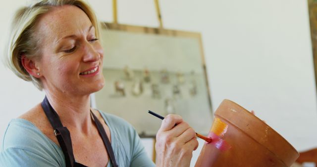 Woman painting a terra cotta pot with a paintbrush. Ideal for depicting creativity, hobbies, crafting activities, and adult engagement in artistic pursuits. Useful for articles and content related to DIY projects, personal hobbies, art-instructional content, and lifestyle blogs focused on creative endeavors.