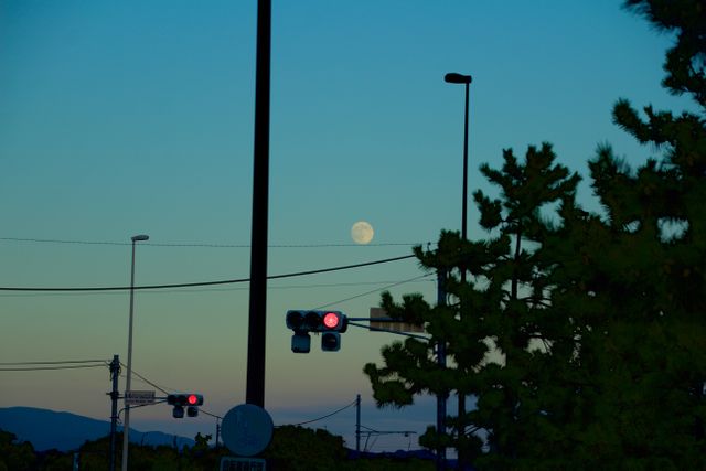 Full moon rising over urban landscape with silhouettes of traffic lights, trees, and lampposts against backdrop of evening sky. Ideal for themes of urban life, city evenings, and ambient lighting.