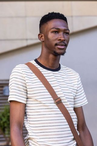 Young man standing outdoors wearing striped tee and brown crossbody strap