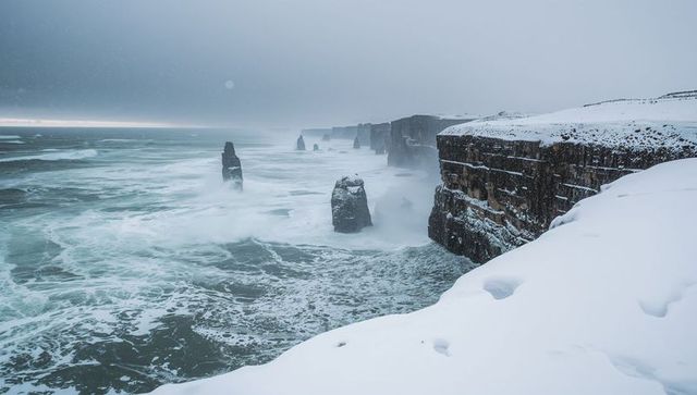 Snow-covered cliffs overlooking stormy teal sea with towering sea stacks in winter
