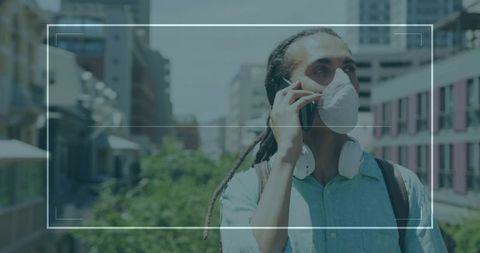 Young man wearing mask talking on phone while walking on urban elevated walkway with headphones and 