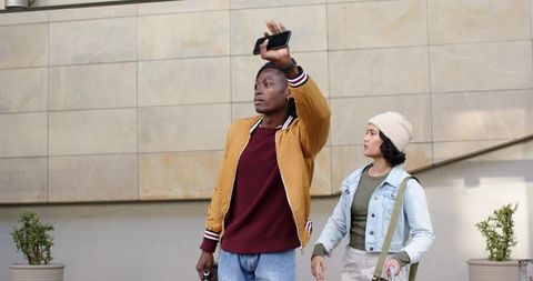 Diverse couple hailing ride with smartphone while pulling luggage in urban area