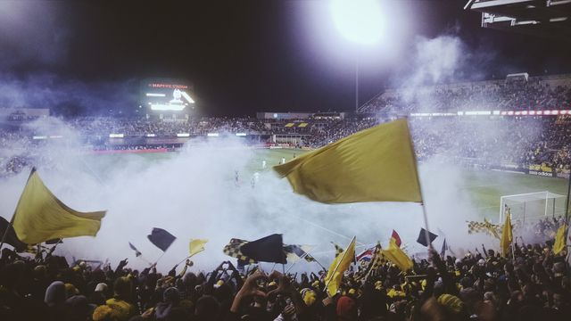 Excited football fans celebrating a goal in a stadium at night with yellow flags and a cloud of smoke. Perfect for use in sports promotions, fan engagement content, and event marketing.