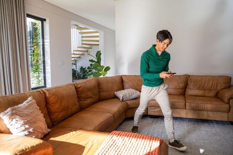 Asian man standing in modern room holding smartphone near leather sectional with plants