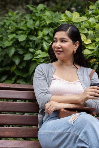 Smiling woman relaxing on park bench holding takeaway cup and leather bag